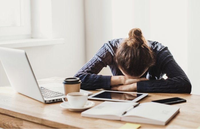 Woman sitting at a desk with her head down on her hands in front of a laptop, coffe cups, tablet, phone, and a book. Blog post featured image.