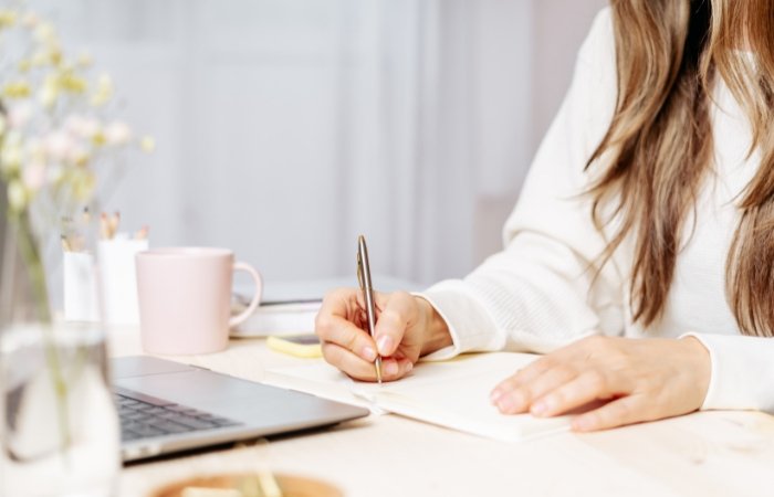 Woman with long hair and a white long-sleeved shirt writing in a book at a desk with a computer, pink mug, and a vase of flowers. Blog post featured image.