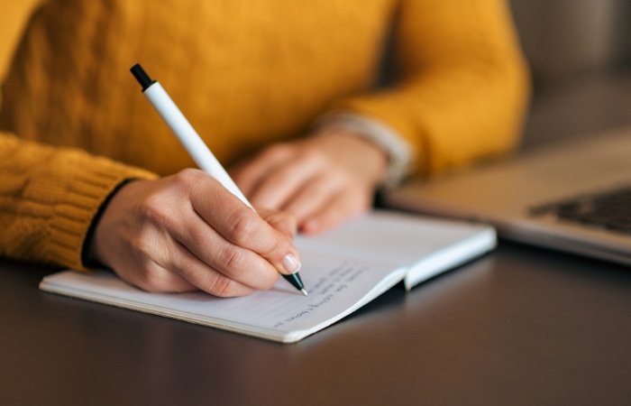 Woman in a yellow sweater sitting at a dark desk writing in a notepad with a laptop open next to her. Blog post featured image.