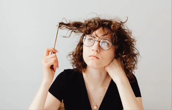 A girl with glasses and brown curly hair is sitting in the center, twirling a pencil around her hair while looking into the distance.